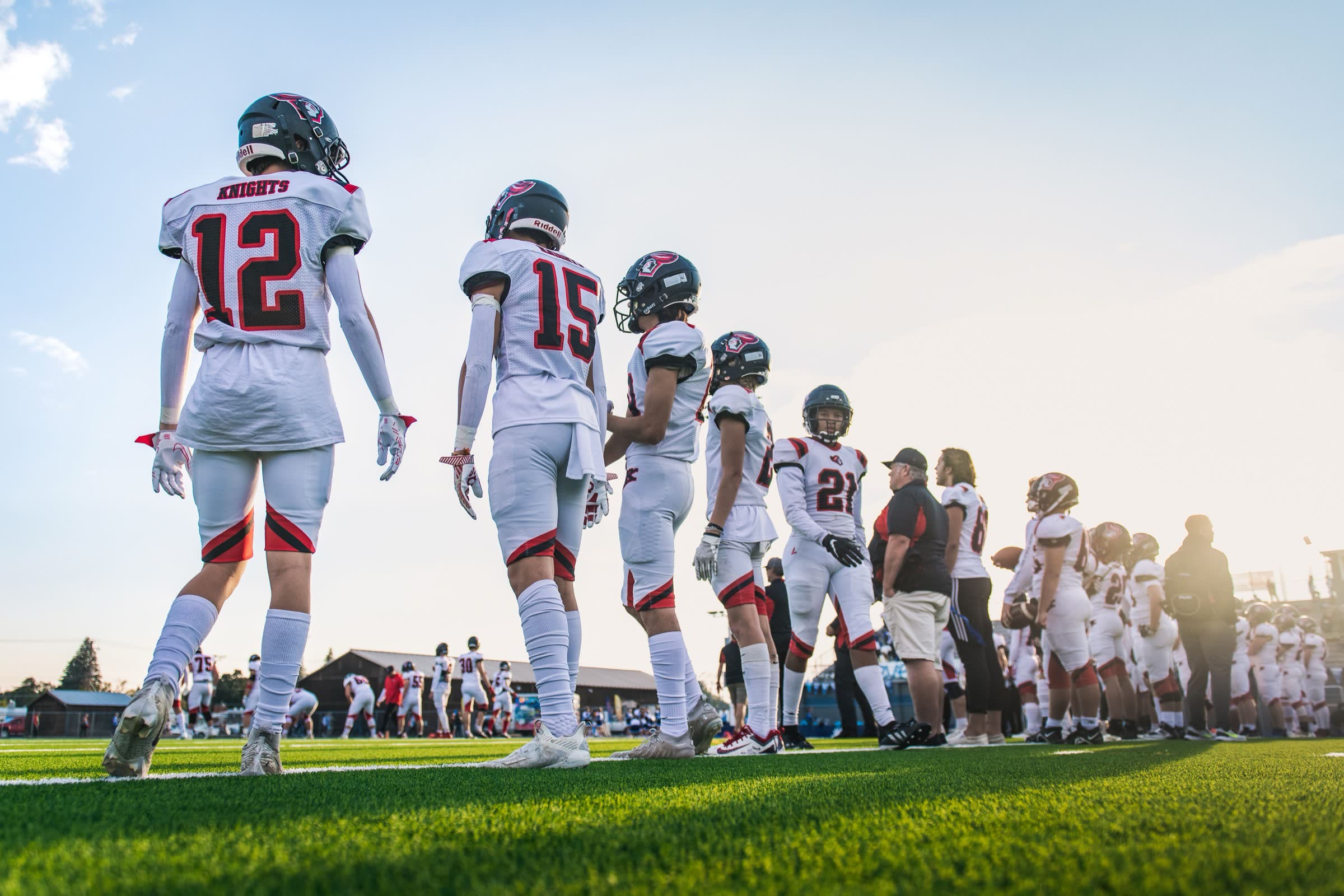 High school football players walking the sideline in low evening sun, teammates and coaches lined along the touchline behind them.
